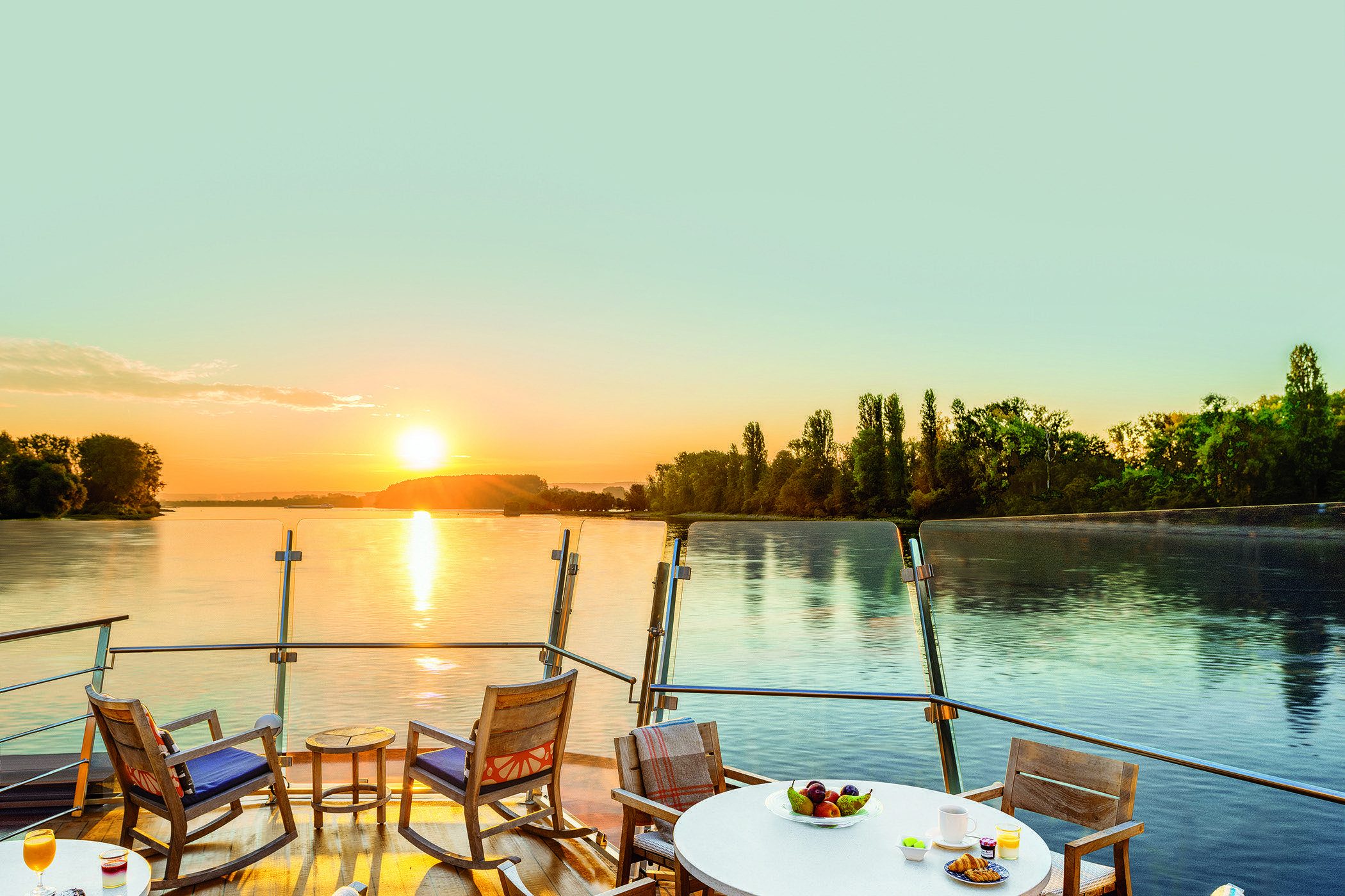 The Aquavit Terrace at sunrise — teak deck chairs with blue cushions and a round table set with fruit and pastries, behind a glass railing, the sun low over a still river.