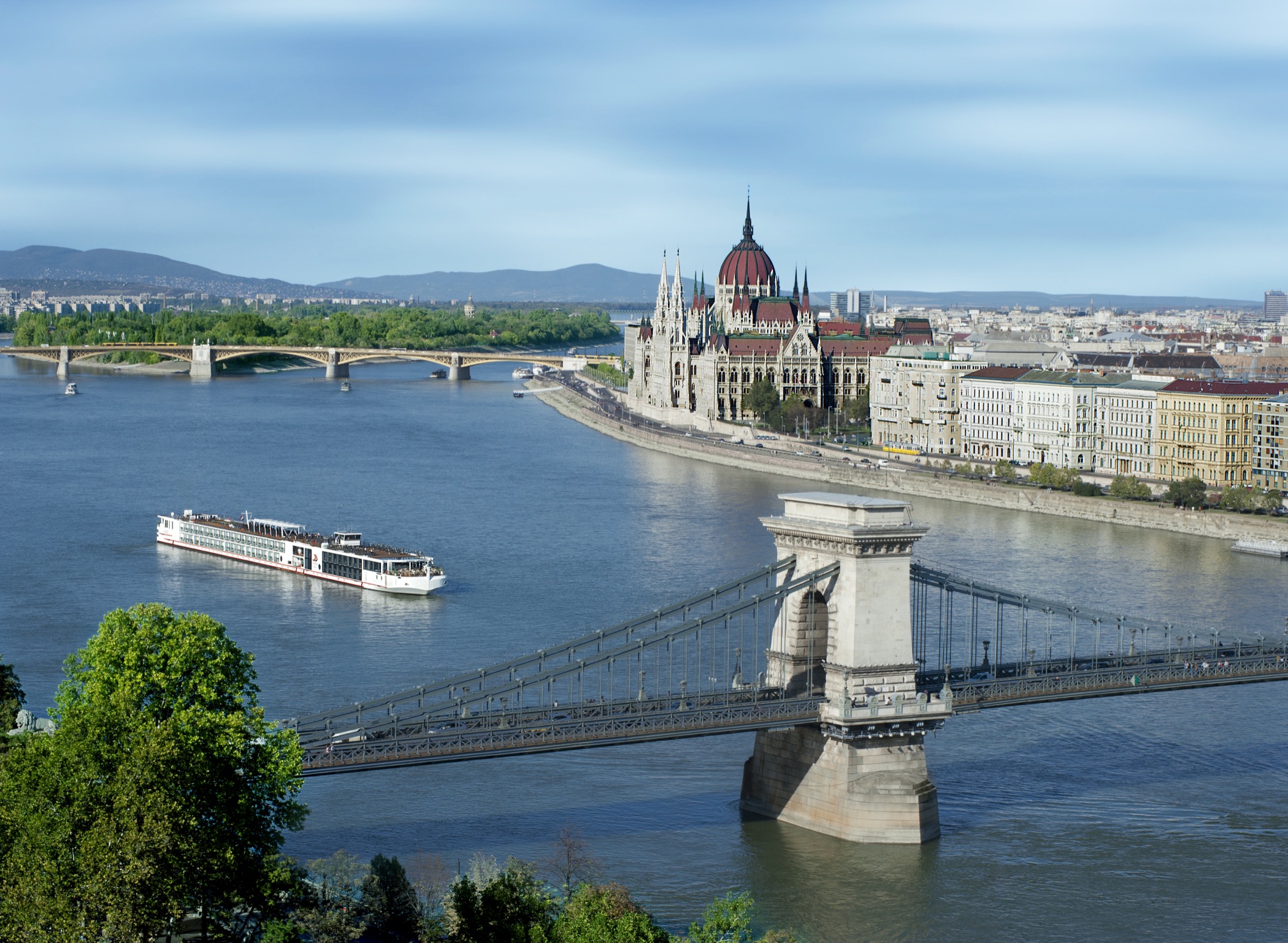 A Viking longship on the Danube in Budapest by day, the Hungarian Parliament and the Pest embankment along the far bank, a tower of the Chain Bridge in the foreground.
