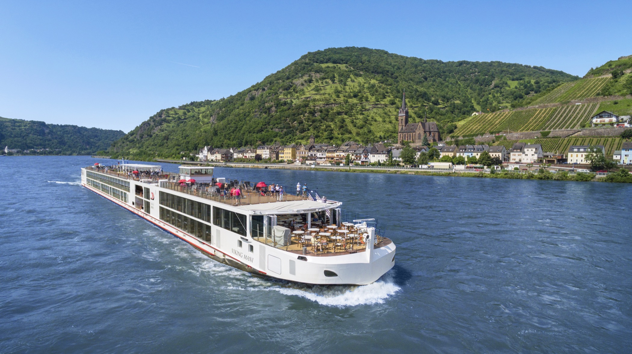 A Viking longship under way on the Middle Rhine, sun umbrellas open on its top deck, passing a village of pale houses and a dark church spire below terraced vineyards.