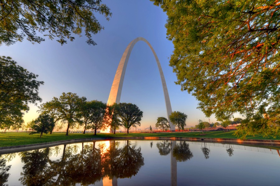 The Gateway Arch in St. Louis rising behind trees and mirrored in a still reflecting pool at sunrise.
