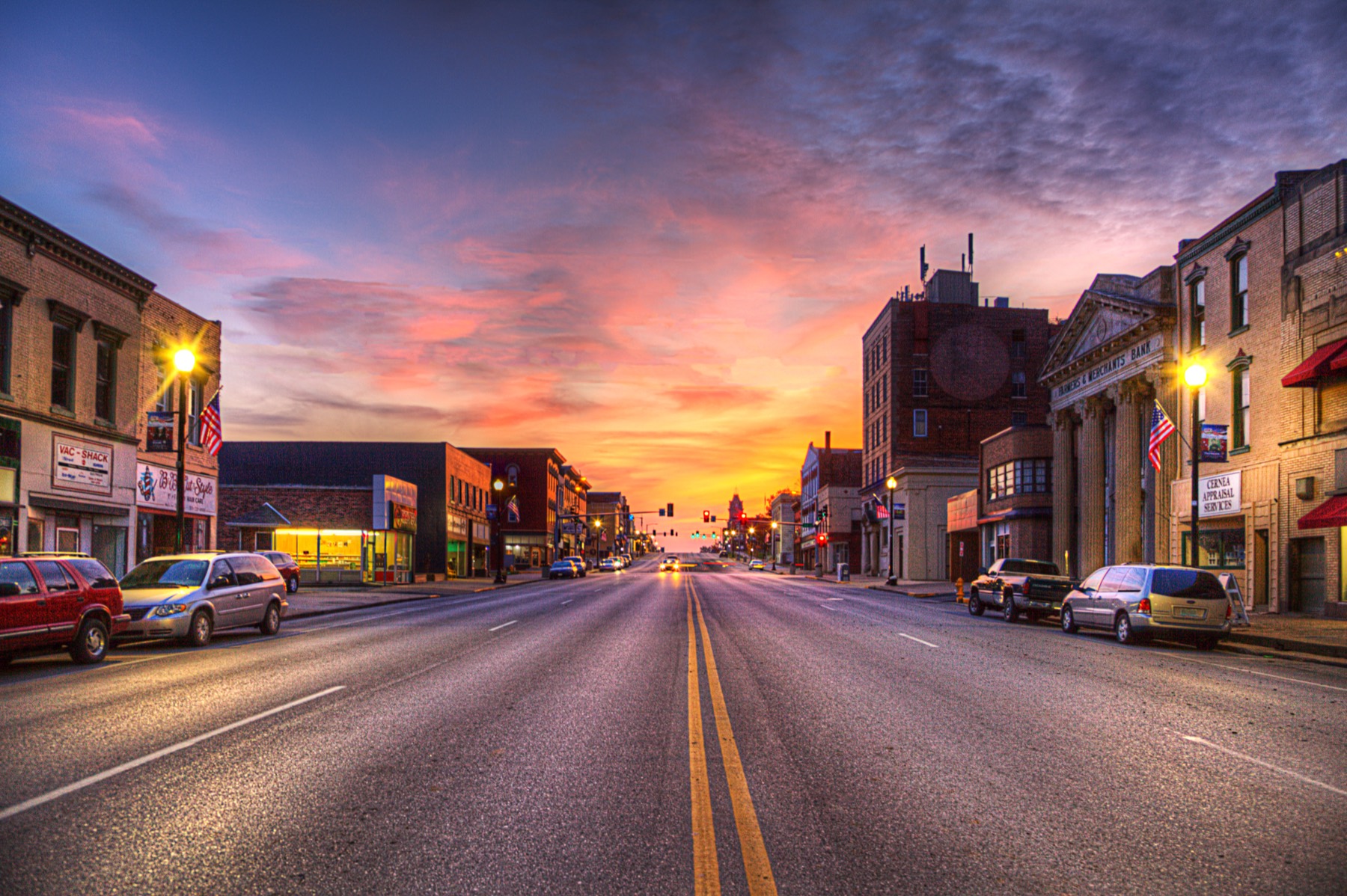 A small river-town main street at sunset, shopfronts and awnings on both sides and the sky burning orange down the length of the road.