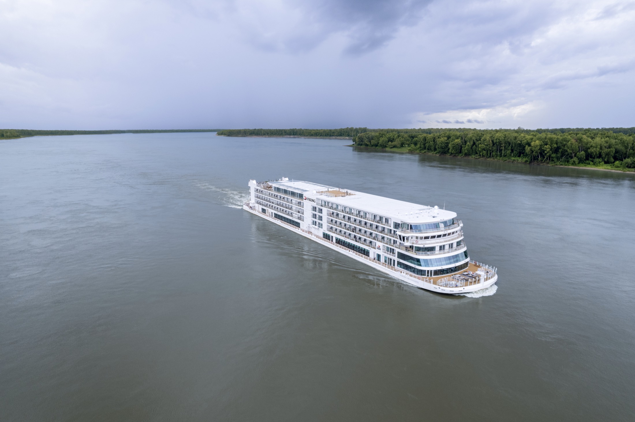 The Viking Mississippi seen from the air, white and low, running down a wide green stretch of the river with wooded banks on either side.