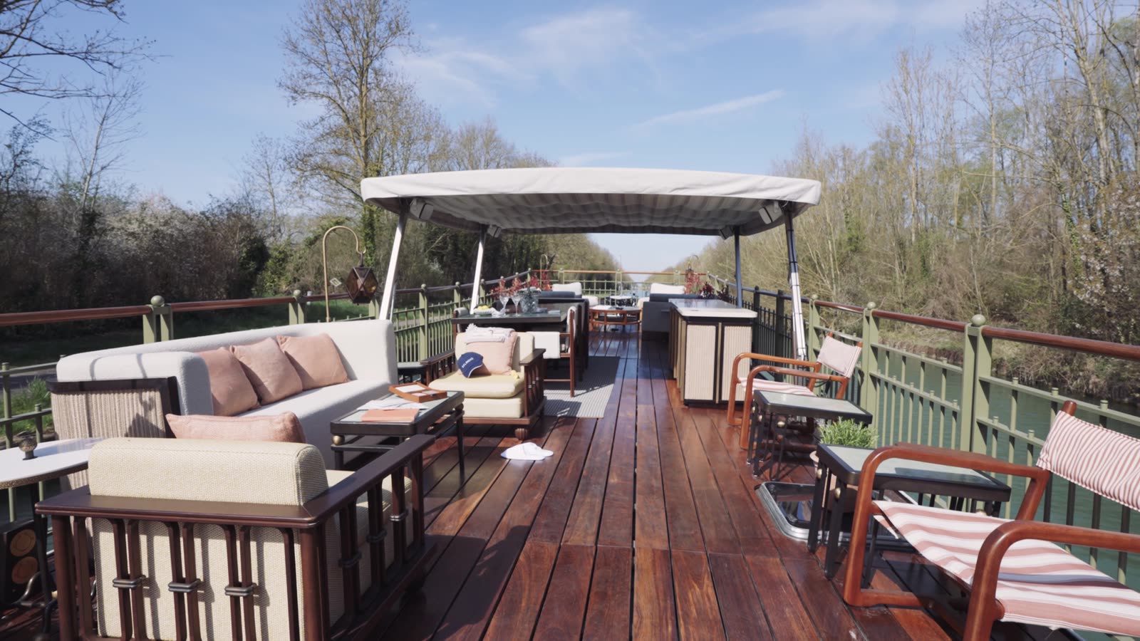 The full upper deck of a Belmond barge moored on a French canal — striped awning, banquette seating, dining table set for evening, hedgerow on the bank.